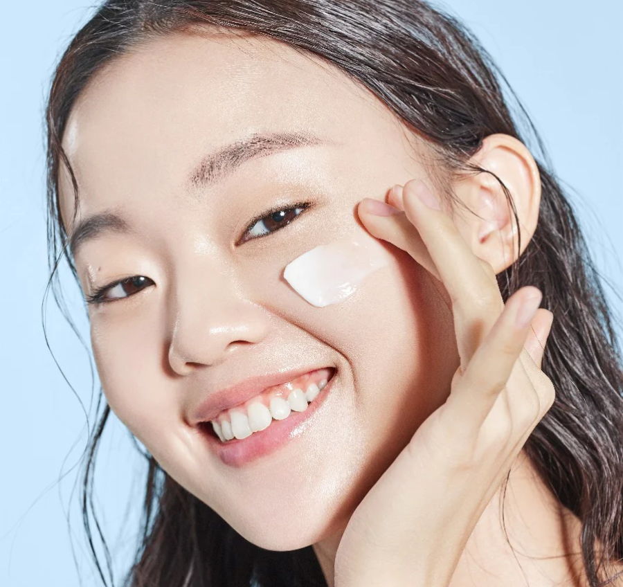 Smiling model applying COSRX The Ceramide Skin Barrier Moisturizer to her cheek, showcasing creamy lightweight texture; Ceramides, Cholesterol, Fatty Acids, Panthenol (B5), and 5 types of Hyaluronic Acid formula locks in hydration, soothes sensitive skin, and enhances glow for smooth, makeup-ready finish.