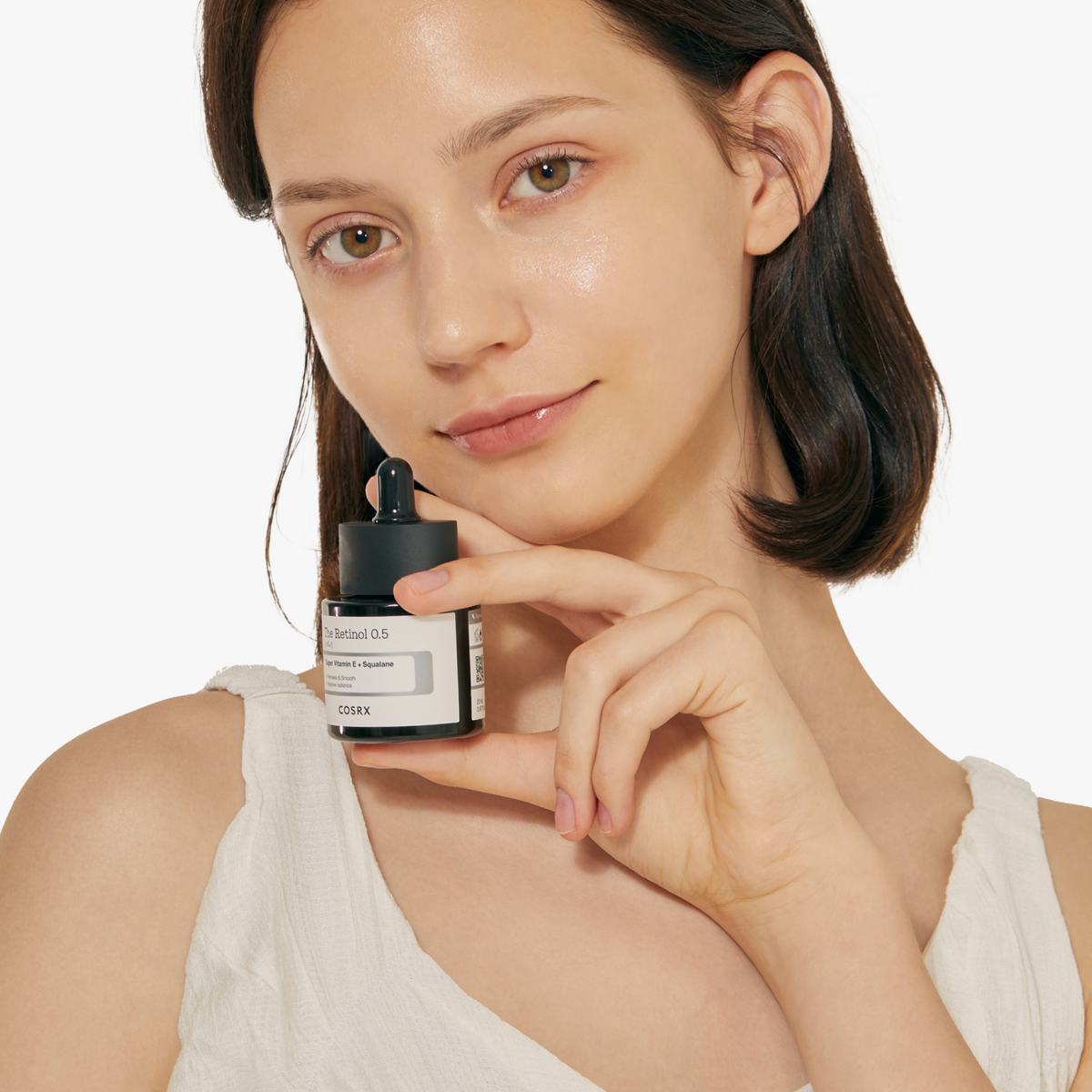 Model with radiant, even-toned skin holding COSRX The Retinol 0.5 Oil close to her face; 0.5% pure stabilized retinol visibly improves wrinkles, fine lines, and skin texture while strengthening the moisture barrier; clinically proven to reduce pore volume by 48.87%, revealing smoother, refined skin with continued use.