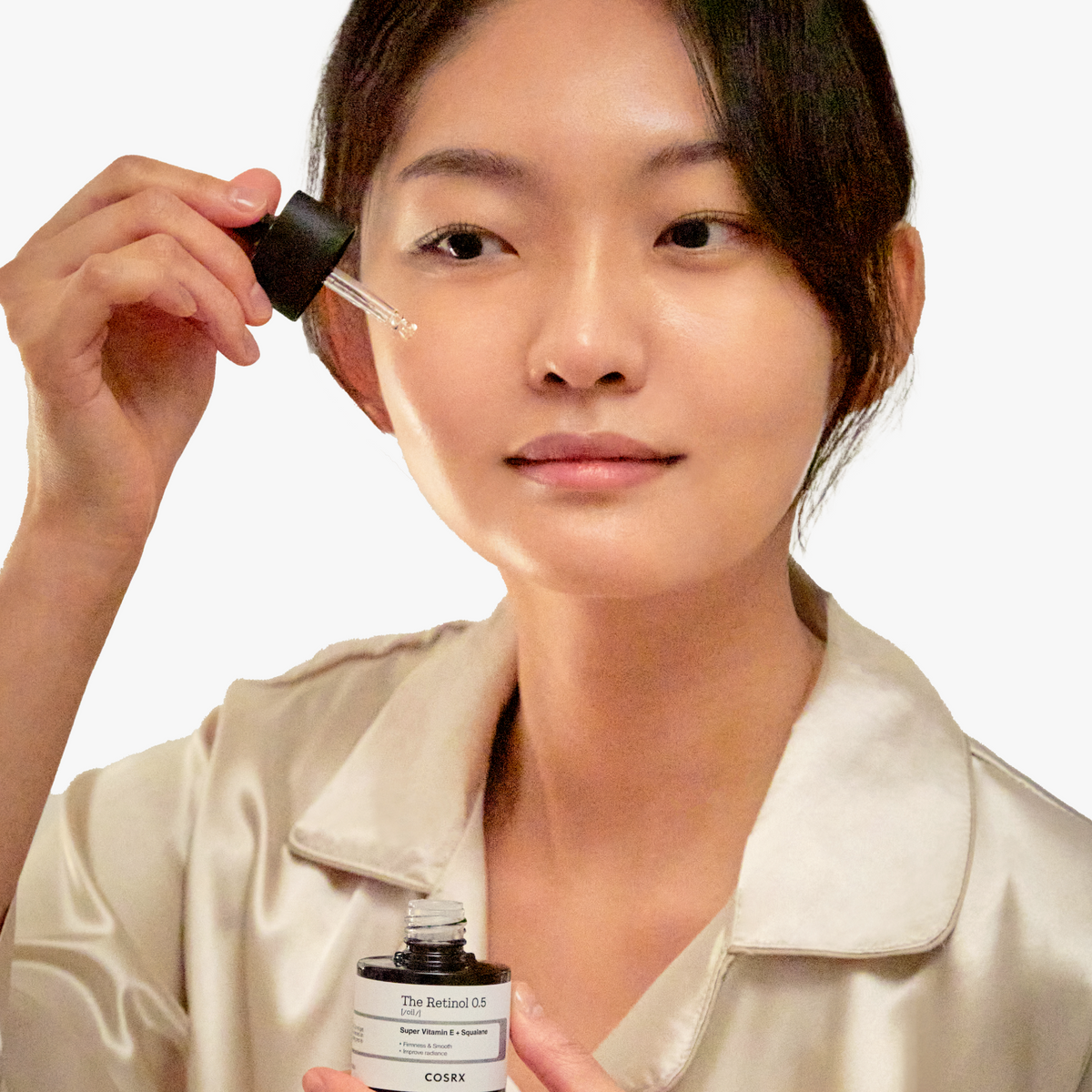 Woman in a silk top applying COSRX The Retinol 0.5 Oil to her cheek with a dropper during a night-time skincare routine; illustrates safe retinol application for intermediate users, promoting wrinkle reduction and elasticity improvement; clinically shown to enhance external skin elasticity by 16.91% after 4 weeks.