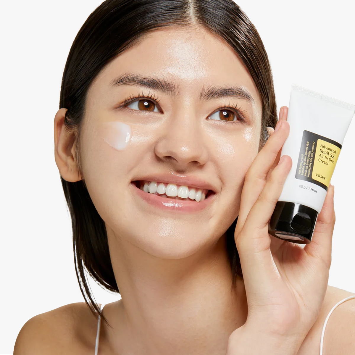 Model smiling while holding COSRX Advanced Snail 92 All In One Cream tube, with cream applied on cheek showing hydrated, luminous skin. Korean moisturizer formulated with 92% snail mucin, clinically shown to reduce transepidermal water loss (TEWL) by 76.46% using the Tewameter, improving barrier strength, elasticity, and lasting glass skin hydration.