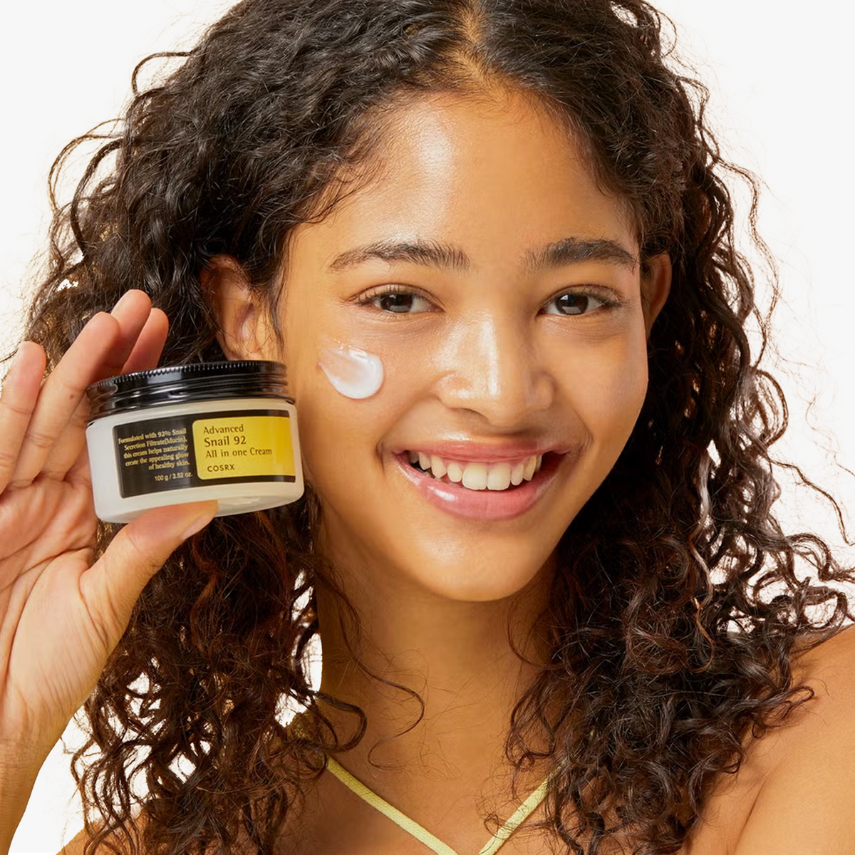 Model with radiant, hydrated complexion holding COSRX Advanced Snail 92 All In One Cream, with cream applied on cheek showing healthy glow and even tone. Formulated with 92% snail mucin, clinically proven to reduce transepidermal water loss (TEWL) by 76.46% using the Tewameter, enhancing skin barrier strength, elasticity, and long-lasting moisture for all skin types.